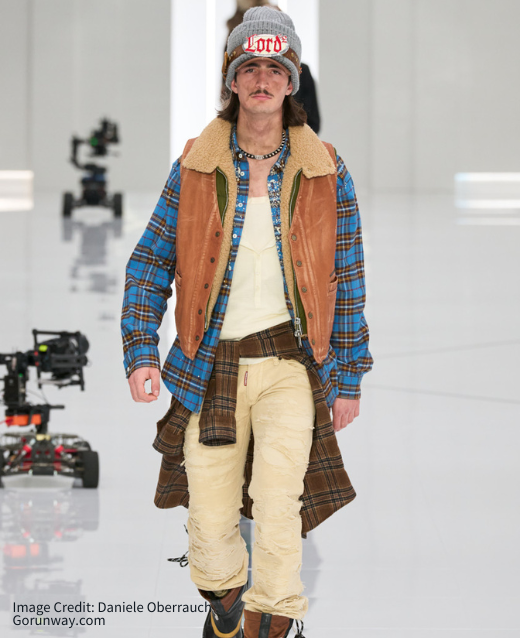 Photo of DSquared2 runway, male model wearing oversized greay beanie with 'Lord' printed in red across it, tartan blue shirt layered with green gilet and brown suede wasitcoat. Another brown tartan shacket is tied around his waist and cream trousers