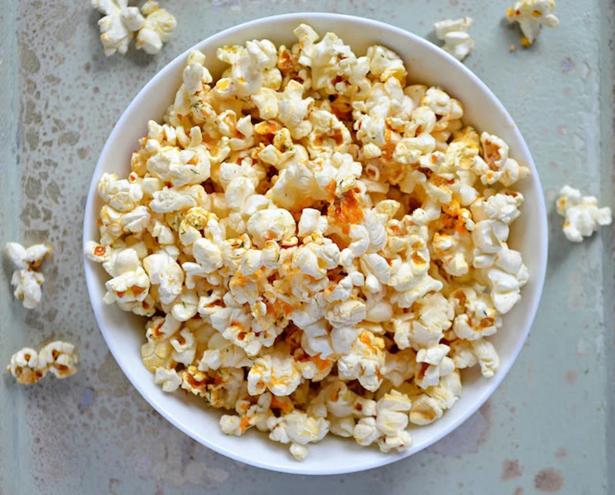 A white bowl filled with Buffalo Ranch Popcorn over flowing onto a light blue counter top.