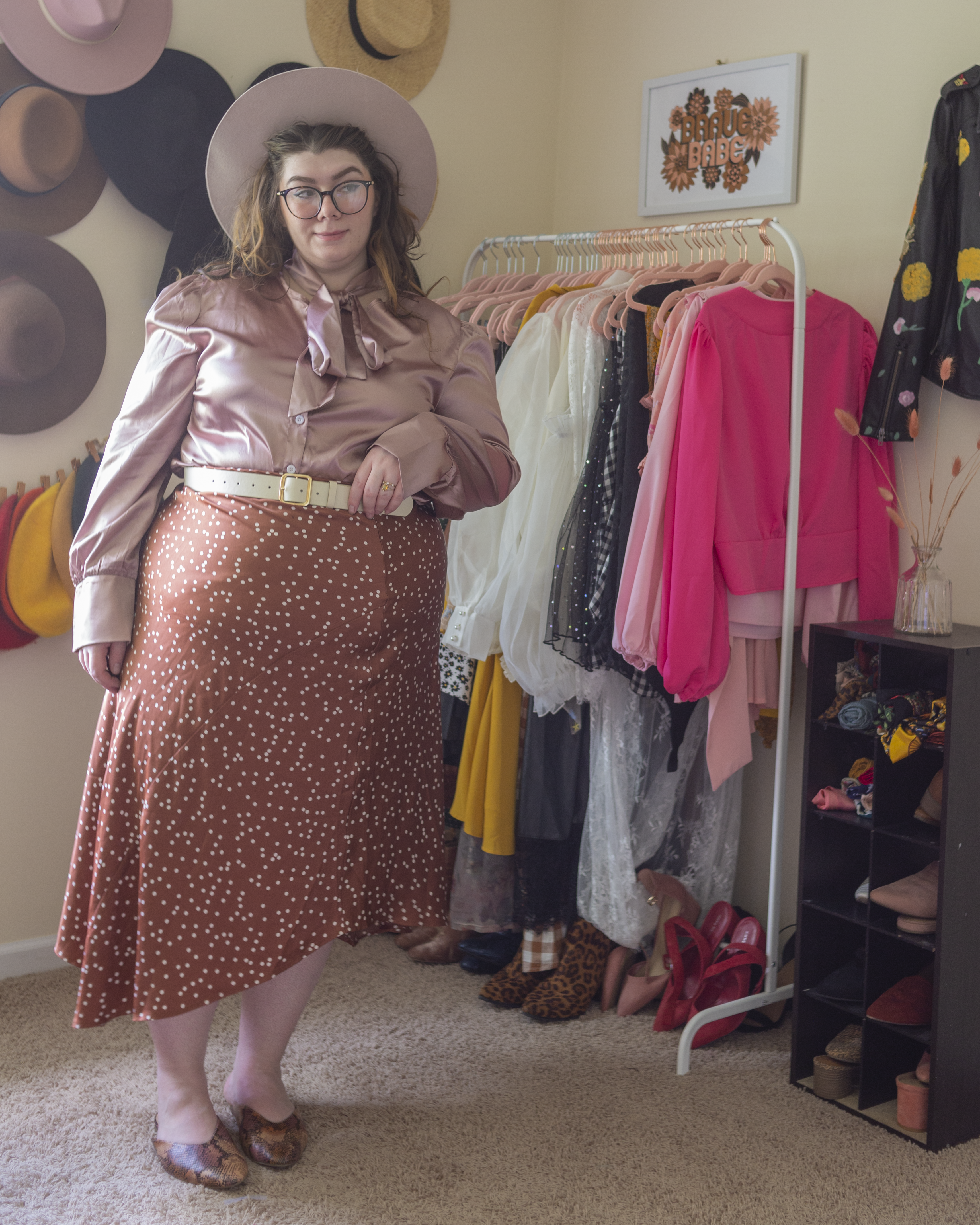 An outfit consisting of a beige and pastel pink pork pie hat, a pastel pink silk button up blouse with an attached pussy bow, tied in a bow around the neck, tucked into a rose pink swiss dot asymmetrical hem slip midi skirt and pastel pink, orange, and brown snake skin mules.