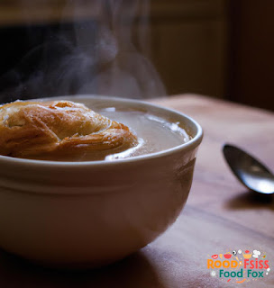 This image shows a close-up of the soup in a bowl with a flaky pastry crust on top. The steam rising from the soup makes it look extra delicious. The bowl is sitting on a wooden table with a spoon nearby.