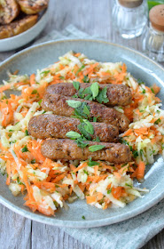 Knusprige gebratene vegane Bratwürstel auf einem Salat aus geraffeltem Kohlrabi und Karotten, bestreut mit Petersilie. Daneben eine Schüssel mit Kümmelkartoffeln.