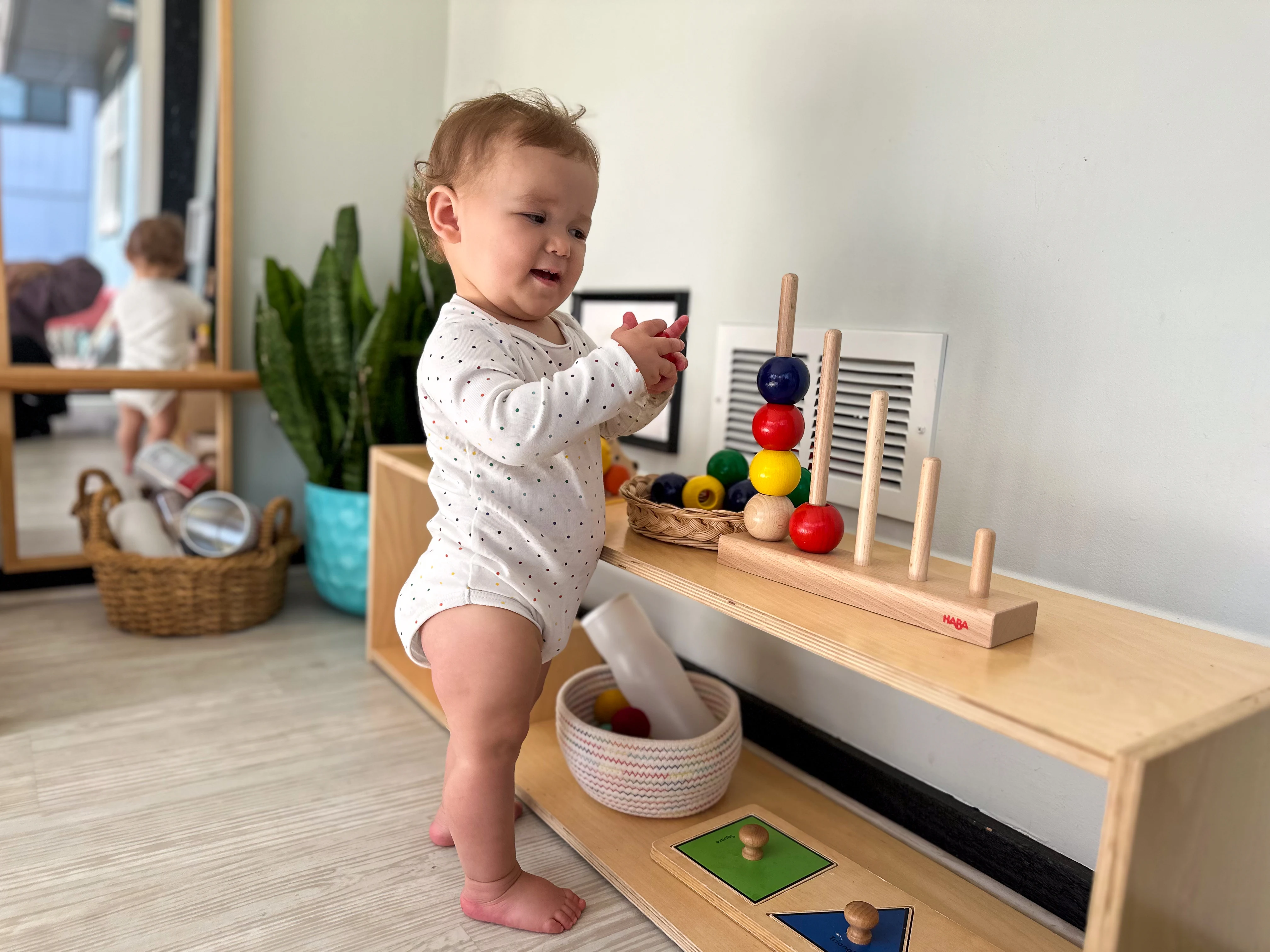 14-month-old Montessori toddler stands at shelf and places colorful balls onto posting toy in her Montessori playroom. These fun toys support Montessori learning at home.
