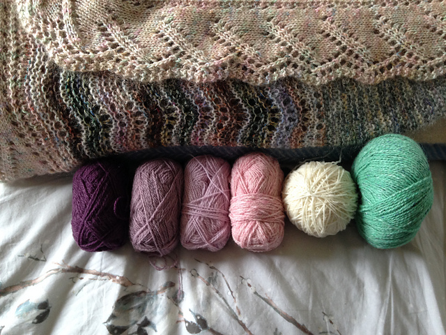 Balls of yarn in colours ranging from deep plum purple, lilac and pale pink to fresh mint green, photographed against a background of hand-knitted blankets neatly folded.