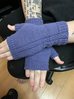 A pair of lavender colored hnadknit fingerless mittens being modeled by a woman with several script tattoos. Only her forearms and hands are visible.