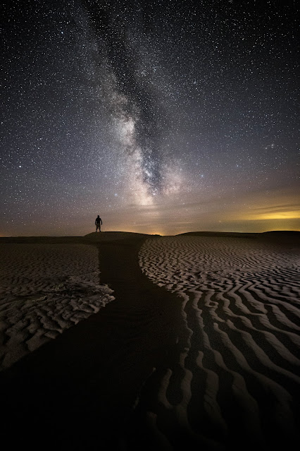 Nighttime image of person on the distant horizon of sandy dunes in small wave pattern under a night sky filled with stars and the milky way