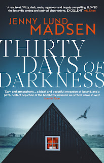 Cover for book "Thirty Days of Darkness" by Jenny Lund Madsen. View over water towards a wintry landscape of low buildings. In the foreground, the triangular gable of a dark, wooden building. In the central of the gable, a single window, it with reddish light, through which is visible a woman working at a laptop.