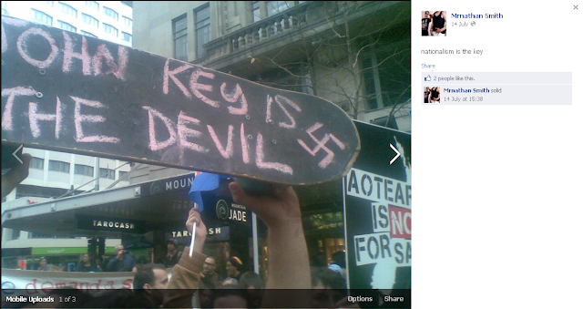 Skate board written in chalk 'John Key is the Devil' with a swastika next to it.  At a protest with an 'Aotearoa is not for sale' sign.