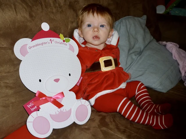 A 3 month old propped up on a sofa in a christmas dress and striped tights next to an oversized  bear shaped card saying "Grandaughters first Christmas"