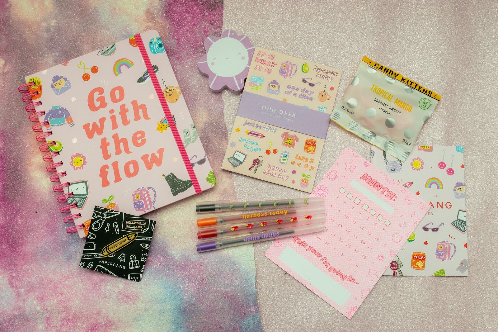 A flat lay of things including a spiral bound notebook, a pin, flower shaped paper notes, stickers, four pens, a postcard and a packet of sweets.