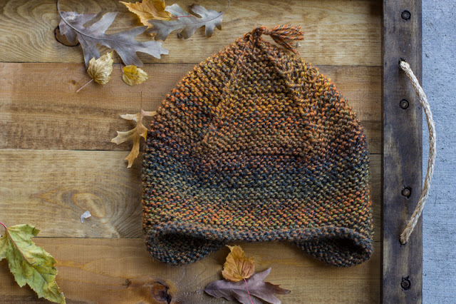 Yellowish, greenish, brownish knit hat with ear flaps and a little tassel coming off the pointed top. Hat lays on a wood tray next to fall leaves.