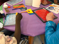 arms and hands shown working on paintings, using paint tools at a table with glue, paint, and tools in the center
