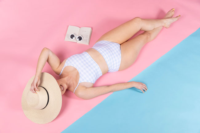 Overhead shot of white woman lying down on pink and blue background wearing pastel blue gingham bikini made with Coralie sewing pattern