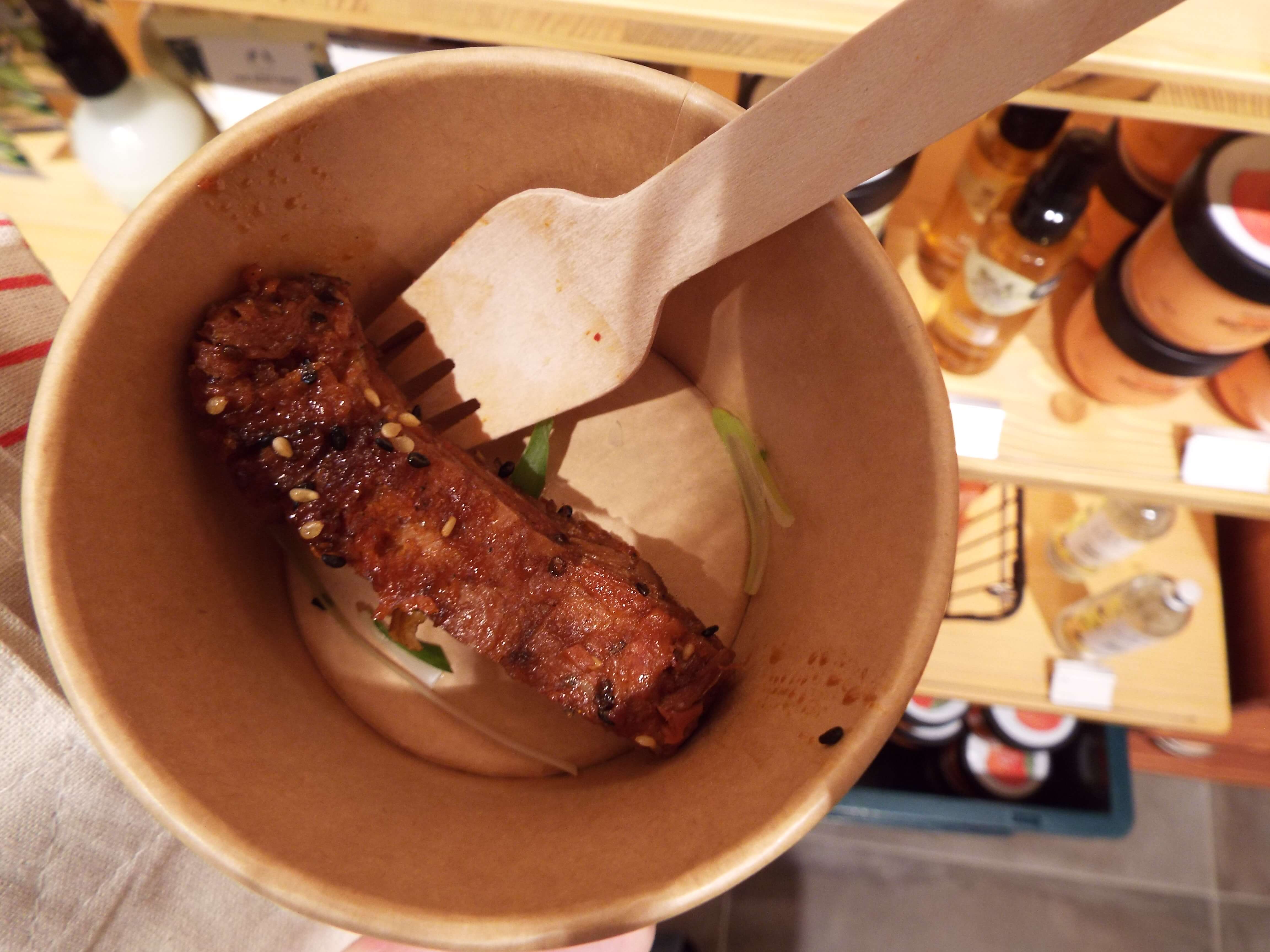 A Birdseye view of the vegan sticky ribs, in a little brown paper tub, seasoned with sesame seeds, with mini wooden fork, amongst the display of beauty products.