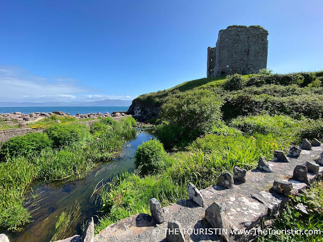 A square castle ruin on a green hill next to a rocky beach and the dark blue ocean under a bright blue sky.