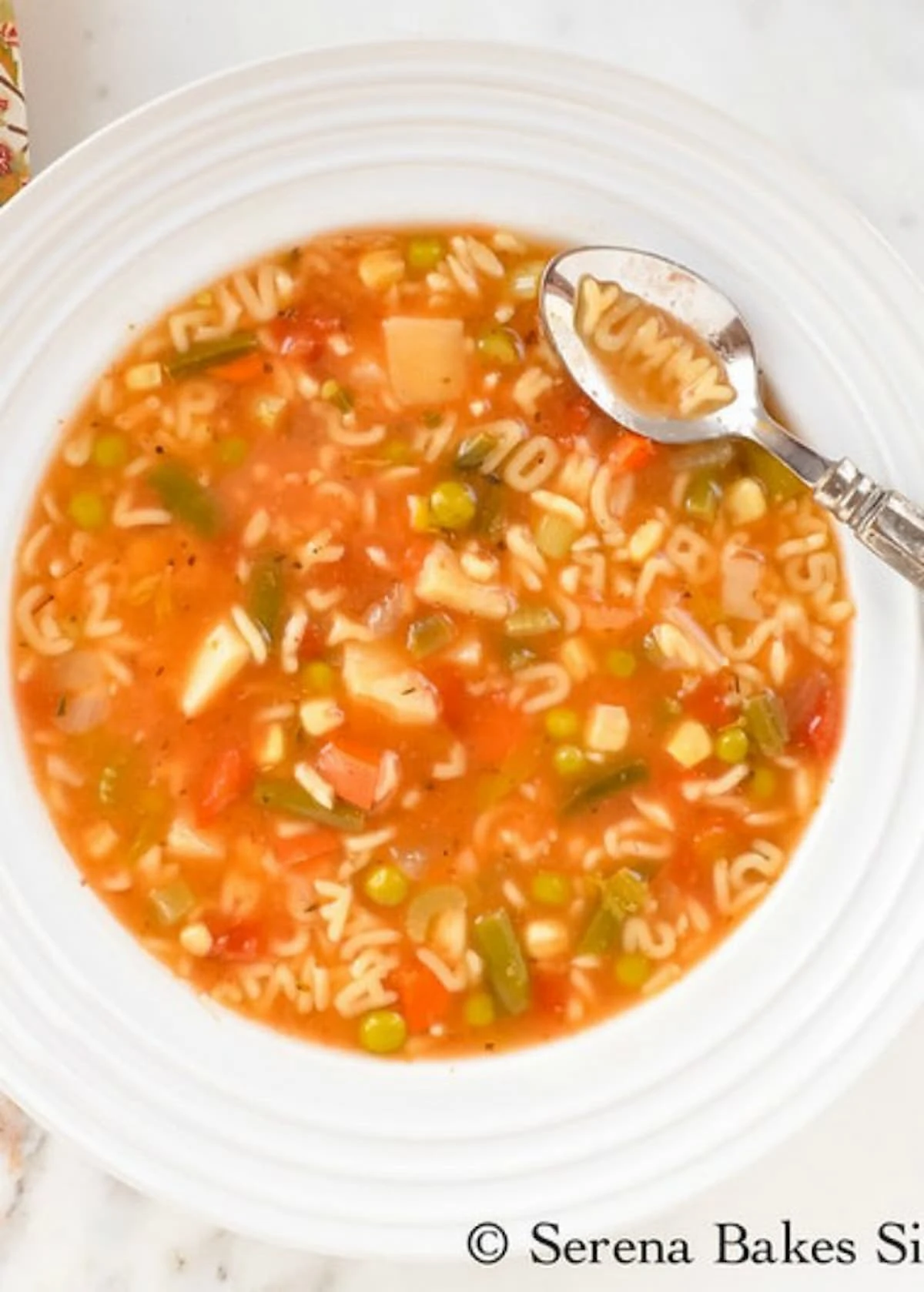 Homemade Alphabet Vegetable Soup filled with alphabet pasta, diced potatoes, green beans, peas, and corn in a white soup bowl with a spoon resting on the edge with the word yummy spelled with alphabet noodles in the spoon.