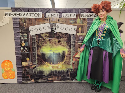 A mural of a cauldron with yellow smoke with the words "Preservation is not just a bunch of Hocus Pocus." A woman stands to the right in a medieval-style dress with purple, black and shades of green. She has a red wig divided into puffs of curls on each side of her head, similar to Bette Midler's character in the film, Hocus Pocus.