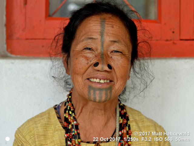 matt hahnewald photography; facing the world; character; face; tattoo; face tattoo; tribal tattoo; eyes; nose; nose plugs; facial expression; eye contact; consent; empathy; emotion; respect; ethnic; traveling; tribal; adivasi; rural; village; traditional; cultural; hong; ziro; arunachal pradesh; northeast india; asian; indian; apatani; one person; female; adult; elderly; woman; picture; photo; illustrative editorial; face perception; physiognomy; educational; nikon d3100; nikkor af-s 50mm f/1.8g; prime lens; 50mm lens; nifty fifty; 4x3 aspect ratio; horizontal orientation; street; portrait; closeup; headshot; seven-eighths view; outdoors; color; posing; authentic; unique; yaping hullo; yesso; shutterstock; smiling; teeth; rapport