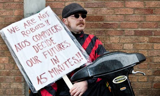 A man wearing dark glasses and seated on a mobility scooter holds a placard reading "We are not robots!! ATOS computers decide our futures in 45 minutes".
