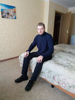 Gleb Zuev is sitting on the bed in trousers, a turtleneck and black Adidas Stan Smith sneakers