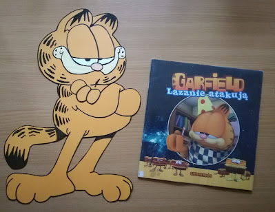 Brązowe tło książka o Garfieldzie i praca plastyczna Garfield stojący z założonymi rękami stworzona na podstawie książek o Garfieldzie