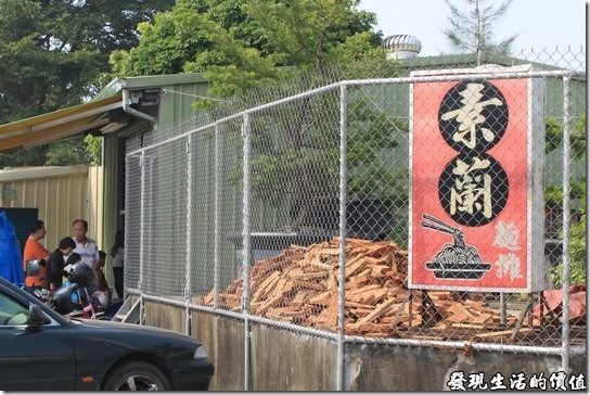 素蘭麵攤。『素蘭麵攤』的招牌，原本擺在下面，為了方便客人停車，所以把招牌移到了上頭，上面的木頭就是用來燒滷汁用的，放在那裡曬太陽，照片中依稀可以看到客人已經排隊排到外面來了。