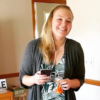 Picture of a blonde-haired, blue-eyed woman (Mandy) wearing a gray sweater and palm tree dress, smiling just off camera while holding a phone and can of seltzer water.