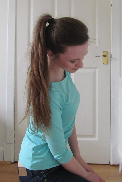 girl with long brown hair in ponytail wearing aqua top sitting next to white door looking down