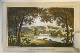 A landscape showing a path running alongside water. There are also cows in the foreground and a bridge in the distance.