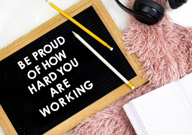 A black sign which says "be proud of how hard you are working". The sign is in a flat lay and has pencils covering one of it's corners. There is also a camera and book in the photo.