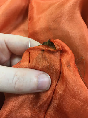 A close-up of a white finger and thumb holding a pinned piece of sheer peach silk backed with green-gold silk. There's a small run in the peach silk just right of the pin.