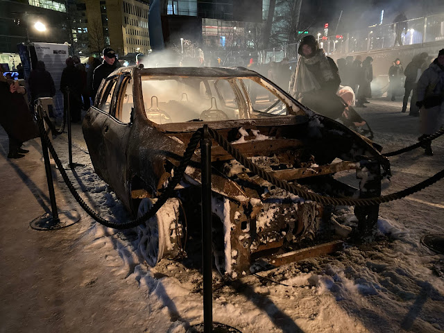 A burnt out car with no wheels has smoke billowing from it, in a snowy, nighttime street