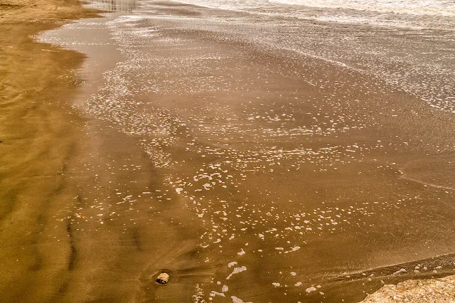 Una piedra en la arena, sola, e primer plano y luego la espuma del mar como acariciando la playa.