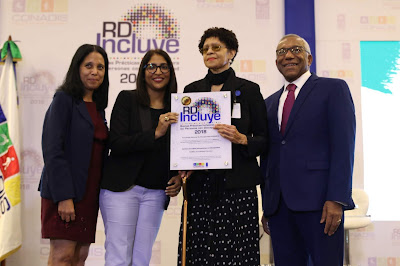 En la foto, de pie, de izquierda a derecha: Maritza Santana del equipo USEPEDI (pelo largo negro, tez clara, cara redonda. Viste chaqueta azul marino, vestido color rojo vino, zapatos negros); Melisa Bretón, miembro del equipo PNUD-RD (pelo largo negro, tez clara, cara alargada, gafas recetadas. Viste chaqueta negra, pantalón lila, zapatos negros); Arlene Severino, encargada USEPEDI (pelo corto castaño rizado, tez clara, cara ovalada con gafas recetadas. Viste bufanda y chaqueta negra, blusa rosada, falda larga, negra con círculos blancos, zapatos negros), sostiene la placa junto con Melisa Bretón; Licdo. Magino Corporán Lorenzo, director del CONADIS (pelo cano, tez oscura, cara redonda, gafas recetadas, chaqueta y pantalón azul marino, corbata roja, camisa blanca, zapatos negros). Todos lucen una amplia sonrisa. Al fondo del escenario una réplica del sello que dice: RD Incluye. Buenas Prácticas Inclusivas para las Personas con Discapacidad 2018