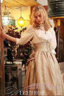 A girl in a tan full, bustled skirt and white shirt with a simple jabot stands in a steampunk tent