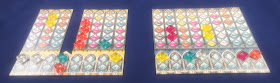 Two player setups. The palace board is wide enough that eight pattern strips can be arranged side-by-side, fitting into the notches along the top.The pattern strips are just wide enough to fit a single pane piece, but tall enough to hold five of them in a column. The palace board is tall enough to hold two pane pieces. Thus, when fully assembled, the pattern strips and pane pieces can hold a maximum of fifty-six pane pieces. The arrangement for the player on the left has one of the pattern strips removed, leaving a gap with three pattern strips on the left side of the palace board, and four on the right side.