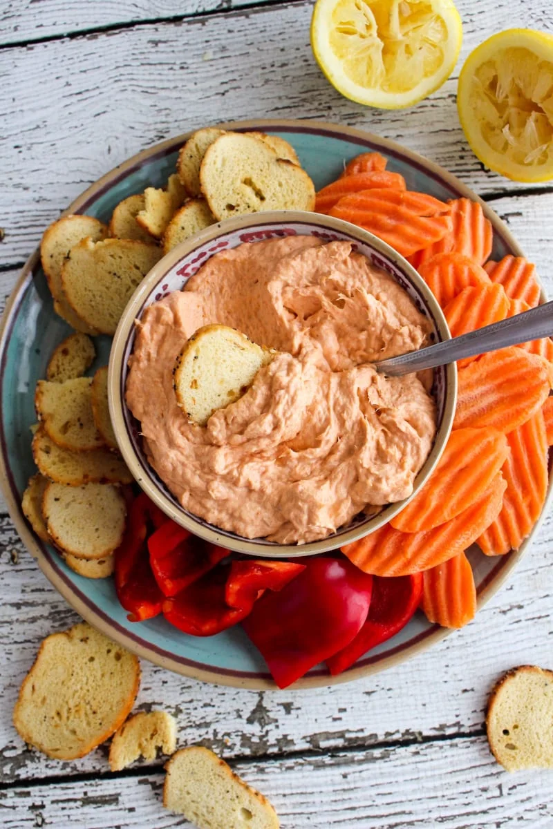 Top view of Smoked Salmon Dip in a tan bowl on a blue plate with bagel chips, carrot chips, and bell pepper pieces around it.