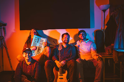 Four musicians sitting in front a wall with a black screen behind them, lit up with an orange and blue light. On the left, the keyboardist holds his electric keyboard, with his head titlted back.