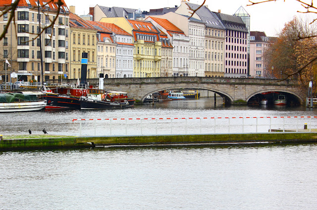 Widok na Sprewę i ulicę Maerkisches Ufer z przeciwległego brzegu rzeki, z Rolandufer. Przy brzegu zacumowane stoją statki, stateczki. Kamienny most biegnie ku brzegom wyspy. Za mostem wznoszą się kamienice, pomalowane w różne kolory, o dachach spadzistych, krytych to czerwoną, to czarną dachówką.  