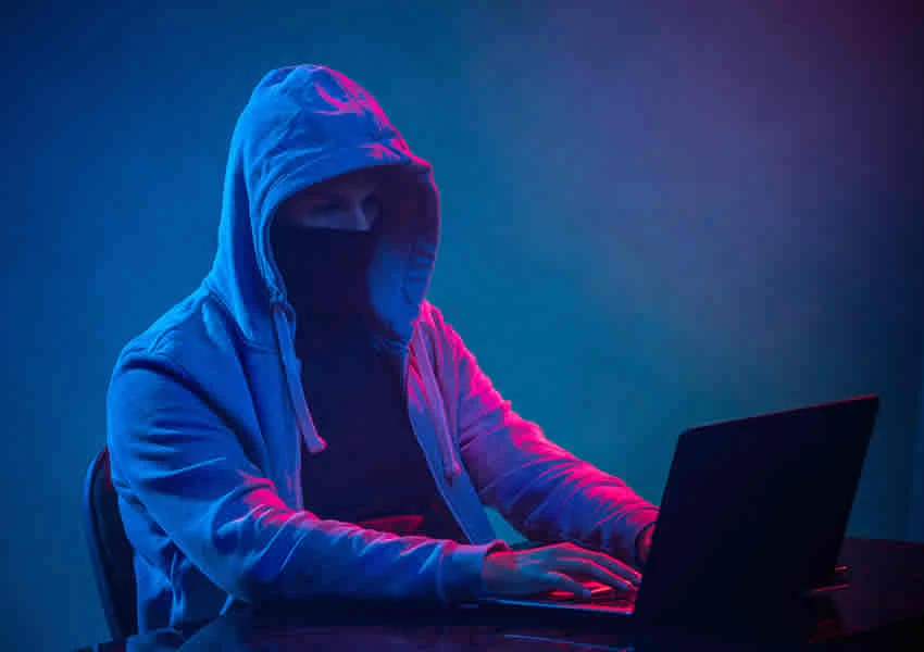 Imagem com fundo azul escuro mostra uma pessoa vestida de capuz manuseando um computador.