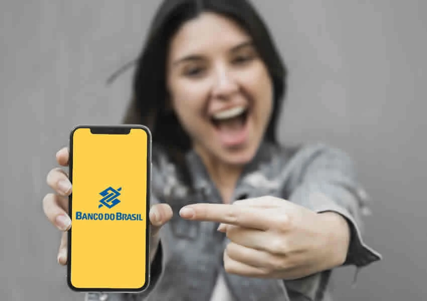 Imagem mostra uma mulher sorrindo segurando com uma das mãos um smartphone e apontando com a outra mão o app do banco do brasil.