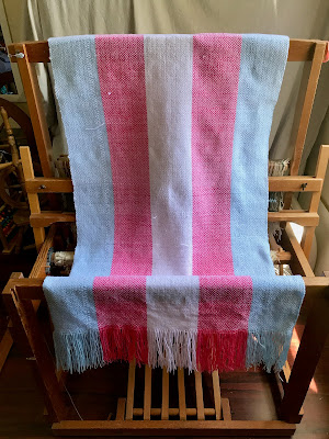 a large piece of fabric is draped over a floor loom. The warp is in the colours of the trans pride flag with white diamonds woven into it. A fringe is visible on the front edge of the fabric, and there are several small white tails of weft thread sticking out of the cloth.