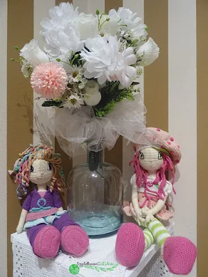 Na stole z białą serwetą, na tle ściany w biało brązowe pasy siedzą dwie lalki wykonane na szydełku. Pomiędzy nimi stoi butelka z białymi kwiatami. Po lewej stronie niższa, ma kolorowe włosy, fioletową bluzkę, różowo-niebieską spódniczkę, fioletowe getry i różowe buty z niebieskim guziczkiem. Po prawej wyższa, ma różowy beret z czerwonymi koralikami, różowe włosy, białą bluzkę, różową, krótką spódniczkę, nogi w zielono-białe pasy i różowe buty.