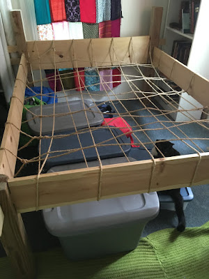 A wooden bedframe with 4 by 4 posts and tall, narrow rails, head, and footboard. Heavy jute rope is tied and woven around the rails to make a rough grid across the frame. A patchwork curtain hangs over the window at the head of the bed, and three grey plastic totes and a black cat sit under the rope lattice.