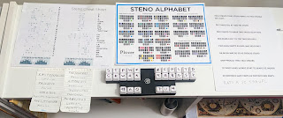 a child's hand pressing a key on a hobbyist keyboard, surrounded by steno reference materials