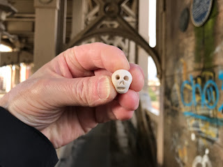 A photo showing a hand holding up a small ceramic skull (Skulferatu 97) with a view of the metal pillars on the lower level of the High Level Bridge in the background.  Photograph by Kevin Nosferatu for The Skulferatu Project.