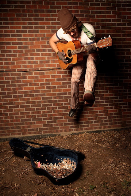 A busker playing guitar and jumping in the air above his guitar case.