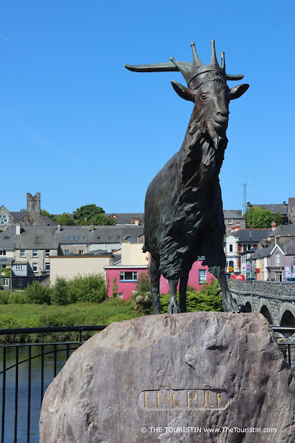 The statue of a goat on a stone in front of a bridge leads to a colourful village with a church..
