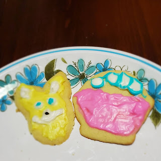 Two cookies sit on a white plate with a blue and green floral border. The left cookie is a deranged rabbit head with yellow fur. The right cookie is a pink Easter basket with white eggs outlined in blue.