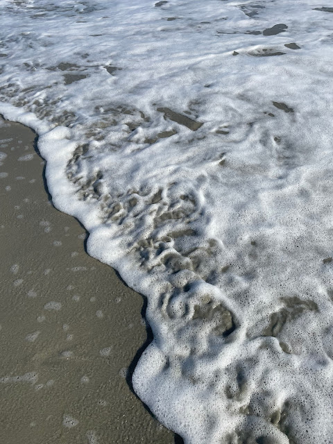 A view of the last part of the wave after it has met the shore. This view shows the line where the wave meets the sand. The water is mostly bubbles and is less than an inch deep. There is nothing else in this picture except for the part of the wave and the sand.