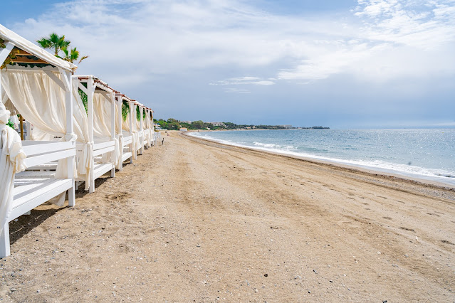 Playa de arena con una línea de pérgolas con cortinas blancas a su espalda y las aguas del mar a su frente.